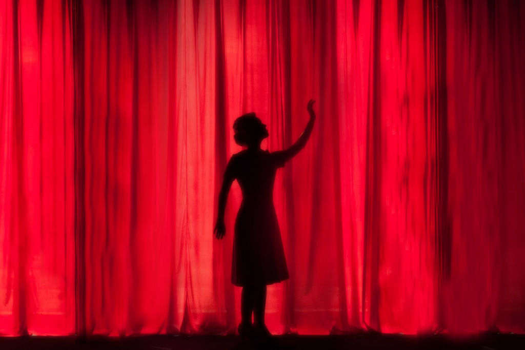 Silhouette of a woman in a dress reaching towards a red curtain, capturing a moment of anticipation before a performance.