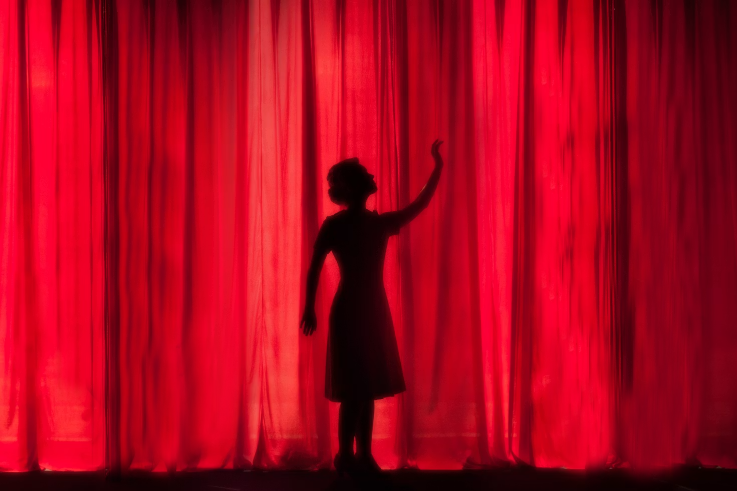 Silhouette of a woman in a dress reaching towards a red curtain, capturing a moment of anticipation before a performance.
